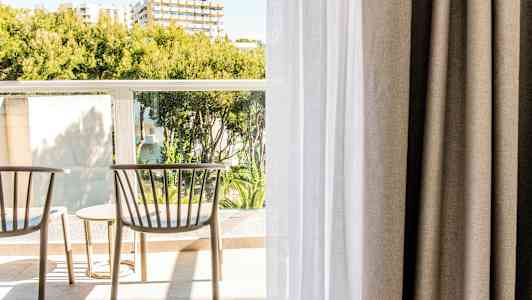 To stole og et bord på en altan med grønne omgivelser og bygninger i baggrunden. (AI-genereret tekst)