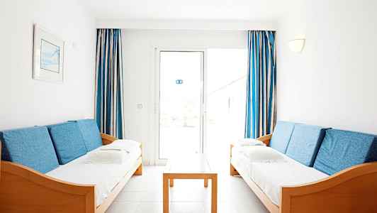 To sofaer med blå puder står over for hinanden, med et træbord imellem og blå-hvide gardiner ved vinduet i baggrunden. (AI-genereret tekst)