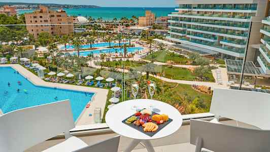 Udsigt fra en balkon mod en hotelpool og havet, med en frugttallerken og to glas på et bord. (AI-genereret tekst)