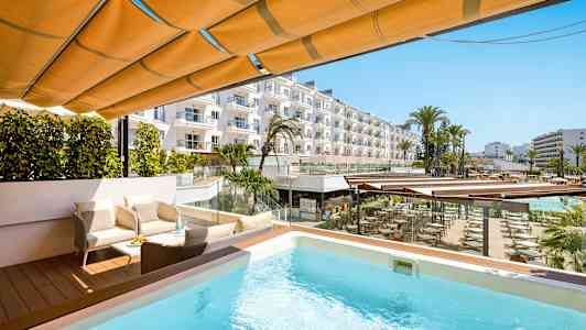 Terrasse med pool, siddegruppe og udsigt over hotelområde med palmer og bygninger. (AI-genereret tekst)