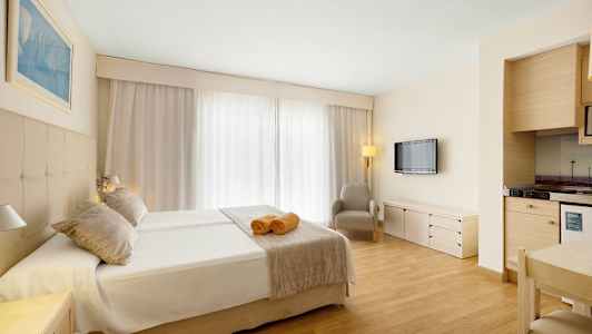 Et hotelværelse med dobbeltseng, to håndklæder på sengen, en lænestol, et vægmonteret TV og et lille køkkenhjørne. (AI-genereret tekst)