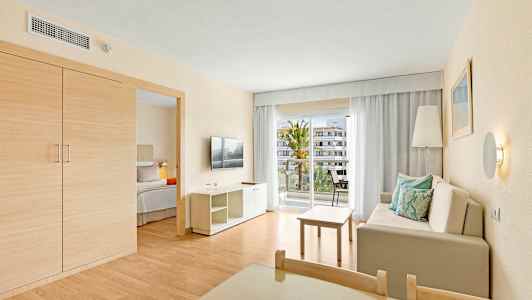 Et hotelværelse med en stue, en sofa med puder, et bord, et TV og en balkon med udsigt. (AI-genereret tekst)