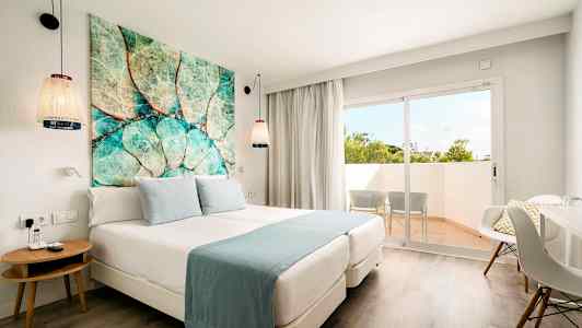 Et hotelværelse med en dobbeltseng, blå sengelinned, et farverigt billede over sengen, og en balkon med to stole udenfor. (AI-genereret tekst)