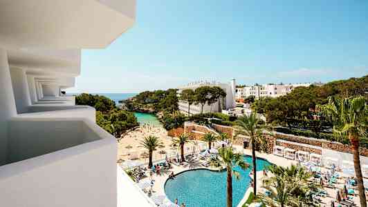 Udsigt fra en altan mod et hotelområde med pool, strand og træer. (AI-genereret tekst)