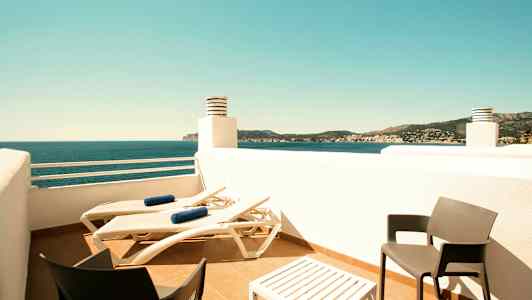 En terrasse med liggestole, et bord og to stole samt udsigt over havet og det omkringliggende landskab. (AI-genereret tekst)