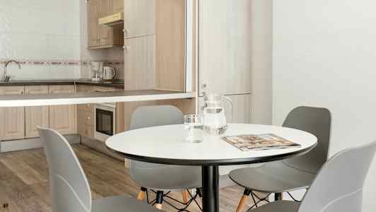 Et spiseområde med et rundt bord, grå stole og et tilstødende køkkenområde med træskabe og hvidevarer. (AI-genereret tekst)