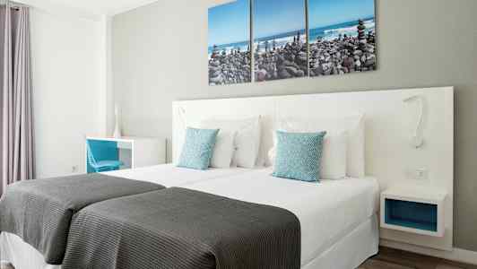 Et hotelværelse med to enkeltsenge, hvidt sengetøj og blå pyntepuder. På væggen hænger et tredelt maleri, der forestiller en stenstrand. (AI-genereret tekst)