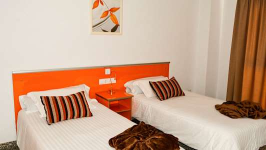 To enkeltsenge med hvide lagner, brune tæpper og stribede puder i et rum med orange interiør. (AI-genereret tekst)