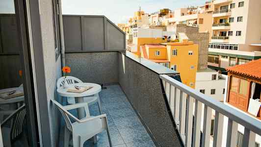 En altan med udsigt til omkringliggende bygninger. Der er et bord med to stole, en bog og en blomst på bordet. (AI-genereret tekst)