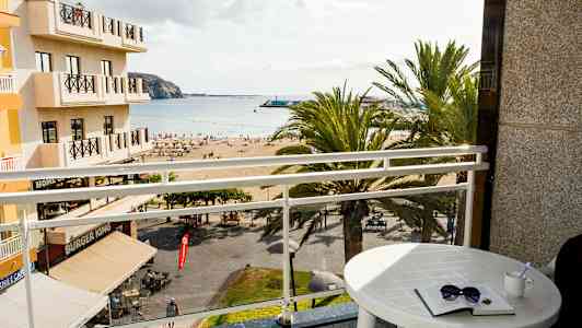 Udsigt fra et hotelværelse med en balkon mod en strand og havet. (AI-genereret tekst)