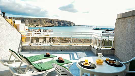 Terrasse med to solvogne og et bord med appelsinjuice og bagværk, havudsigt i baggrunden. (AI-genereret tekst)