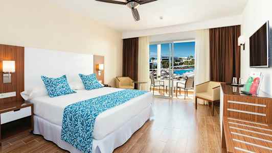 Et hotelværelse med en stor seng, blå dekorative puder og tæppe samt et bord med stole ved en glasdør, der fører til en balkon. (AI-genereret tekst)