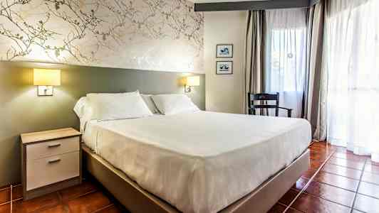 Et værelse med en dobbeltseng, natbord, stol og vindue med gardiner. (AI-genereret tekst)