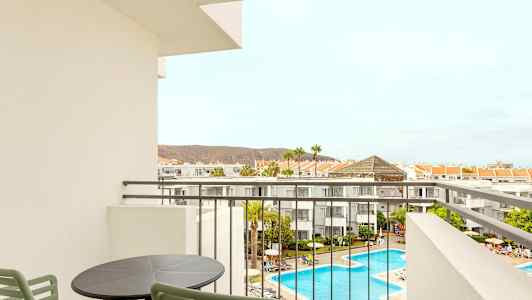 Udsigt fra en balkon over hotelområdet med pool og omkringliggende bygninger. (AI-genereret tekst)