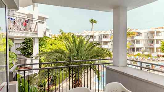 Udsigt fra en altan mod flere hvidmalede bygninger med frodige palmer og et poolområde nedenfor. (AI-genereret tekst)