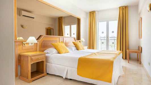 Et hotelværelse med en dobbeltseng, hvide lagner og gule puder og sengetæppe. Værelset har lyse trémøbler og vinduer med gardiner. (AI-genereret tekst)