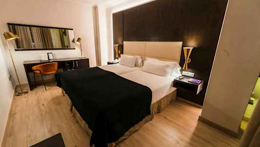 Et hotelværelse med en dobbeltseng, mørke og lyse farvetoner på møbler samt et skrivebordshjørne med stol og spejl. (AI-genereret tekst)