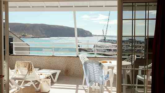 Udsigt fra en altan mod havet og en lille havn, med havemøbler og liggestole på altanen. (AI-genereret tekst)