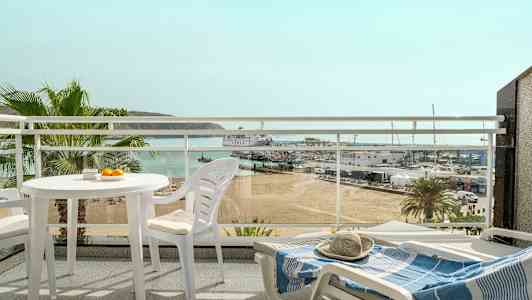 Udsigt fra en altan med hvide havemøbler, med blik over en strand, en lystbådehavn og havet i baggrunden. (AI-genereret tekst)