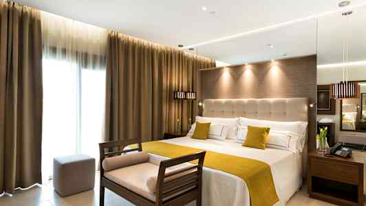 Hotelværelse med stor seng, lyse vægge og gardiner samt en bænk ved fodenden. (AI-genereret tekst)