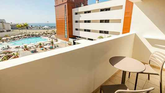 Udsigt fra en altan mod et poolområde med liggestole, parasoller og palmer samt hvide og røde bygninger og havet i baggrunden. To stole og et bord ses på altanen. (AI-genereret tekst)