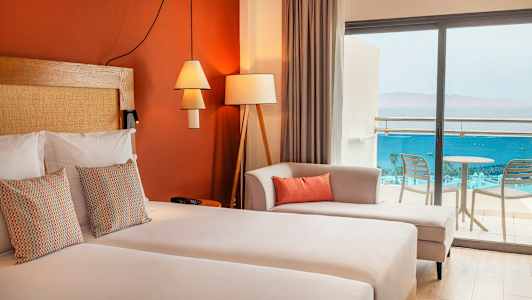 Et hotelværelse med dobbeltseng, dekorative puder, en sofa og glasdøre, der fører til balkonen med udsigt over havet. (AI-genereret tekst)