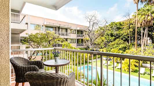 Udsigt fra en altan med to kurvestole ved et lille bord over en pool og grønne områder, samt en boligbygning i baggrunden. (AI-genereret tekst)