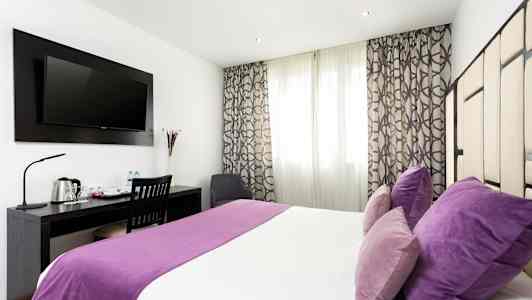 Et hotelværelse med dobbeltseng, lilla sengetøj og puder, skrivebord med stol og en vægmonteret tv. (AI-genereret tekst)