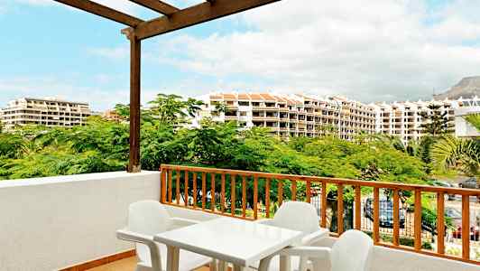 Udsigt fra en altan med plastikbord og stole, omgivet af grønne træer og bygninger i baggrunden. (AI-genereret tekst)