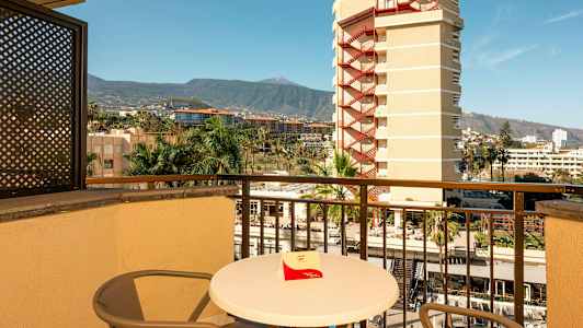 En altan med bord og stole, udsigt over omkringliggende bygninger, palmer og bjerge i baggrunden. (AI-genereret tekst)