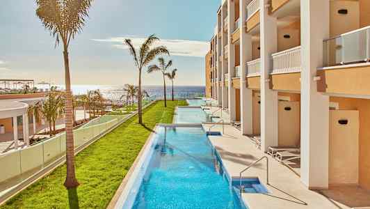 Langstrakt pool omgivet af grønne områder og bygninger med altaner, med havet i baggrunden. (AI-genereret tekst)