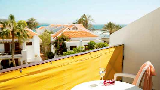 Udsigt fra en altan med gul afskærmning, et lille bord med glas og røde solbriller, hvid stol samt ferieboliger og havet i baggrunden. (AI-genereret tekst)