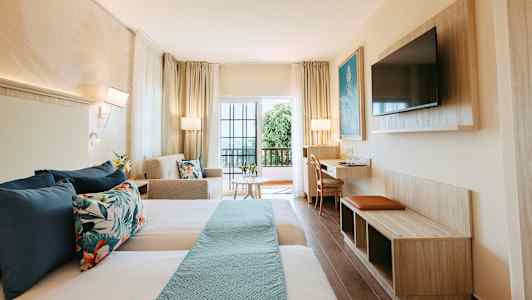 Et hotelværelse med to senge, sofa, bord og TV samt en altan med glasdøre. (AI-genereret tekst)