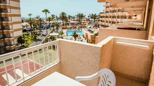 Udsigt fra en altan mod en pool og omkringliggende bygninger i et solrigt område. (AI-genereret tekst)