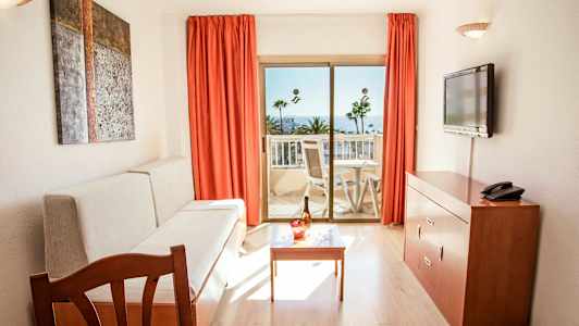 Værelse med sofa, bord, TV og altan med udsigt til palmer og hav. (AI-genereret tekst)