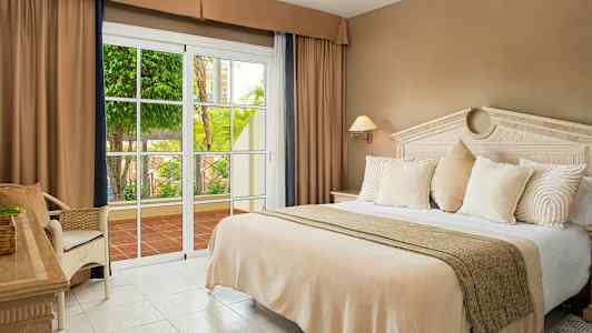Et hotelværelse med en stor seng, beige sengetøj, hvide puder og en glasdør, der åbner ud til en terrasse med beplantning. (AI-genereret tekst)