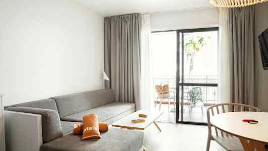 Et hotelværelse med en grå siddegruppe, et bord med træstole og en terrasse gennem glasdøre. (AI-genereret tekst)