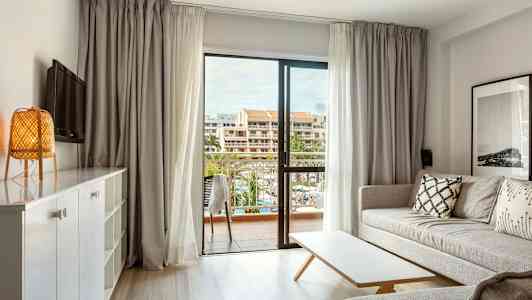 Lyst opholdsrum med sofa, bord og udsigt til en altan med bygninger i baggrunden. (AI-genereret tekst)