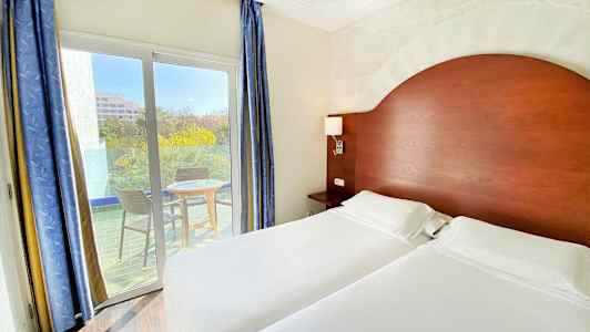 Et hotelværelse med to enkeltsenge, et træhovedgærde og en balkon med to stole og et bord udenfor. (AI-genereret tekst)