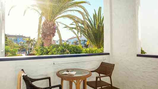 En balkon med to stole og et bord med en bog. Bag balkonen ses en palme og bygninger. (AI-genereret tekst)