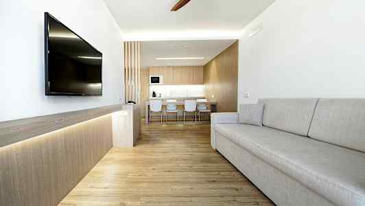 En moderne stue med lys indretning, en grå sofa, et vægmonteret TV og et køkken i baggrunden. (AI-genereret tekst)