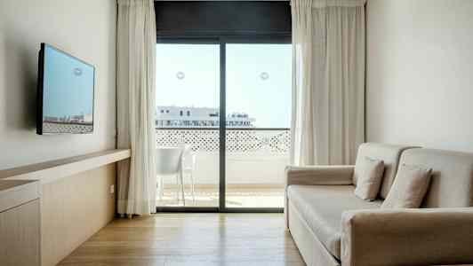 Et lyst og moderne hotelværelse med sofa, vægmonteret TV og stort vindue med udsigt til en altan. (AI-genereret tekst)