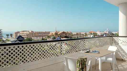 Udsigt fra en altan med bord og stole over et bymiljø med havet i baggrunden. (AI-genereret tekst)