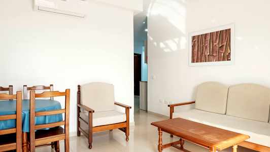 Et rum med lyse vægge og træmøbler, herunder et spisebord med stole, en lænestol og en sofa. (AI-genereret tekst)