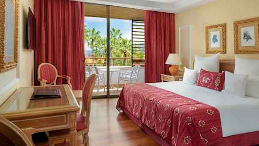 Et hotelværelse med en stor seng, sidemøbler og en altan med udsigt til palmer. (AI-genereret tekst)