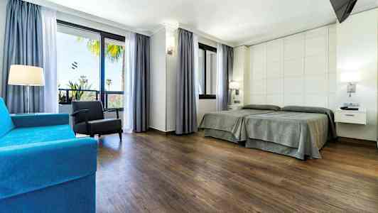 Et hotelværelse med to enkeltsenge, en blå sofa, en lænestol og store vinduer med udsigt. (AI-genereret tekst)