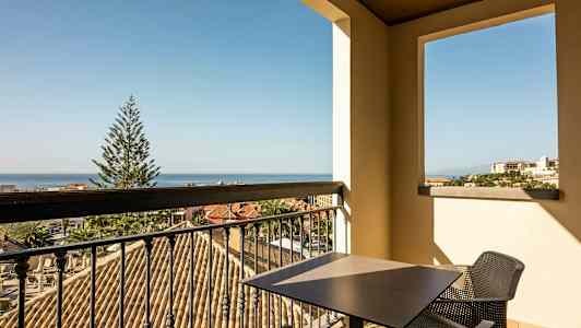 Udsigt fra en balkon med bord og stole over et taglandskab, et træ og havet i baggrunden. (AI-genereret tekst)