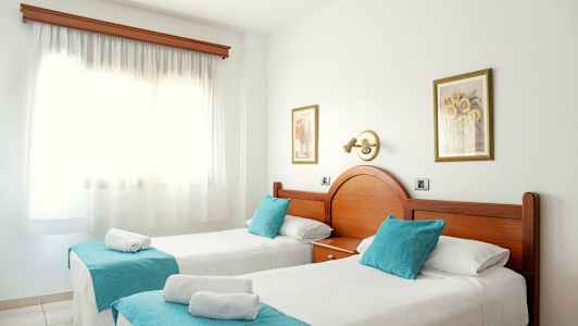 To enkeltsenge med turkise pyntpuder og hvidt sengetæppe i et solrigt rum med et stort vindue. (AI-genereret tekst)