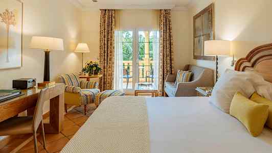 Hotelværelse med seng, skrivebord og siddeområde foran vindue, der fører til altan. (AI-genereret tekst)