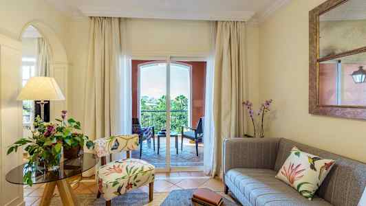 En stue med dekorativ stol, sofa og blomster, med udsigt til en balkon. (AI-genereret tekst)